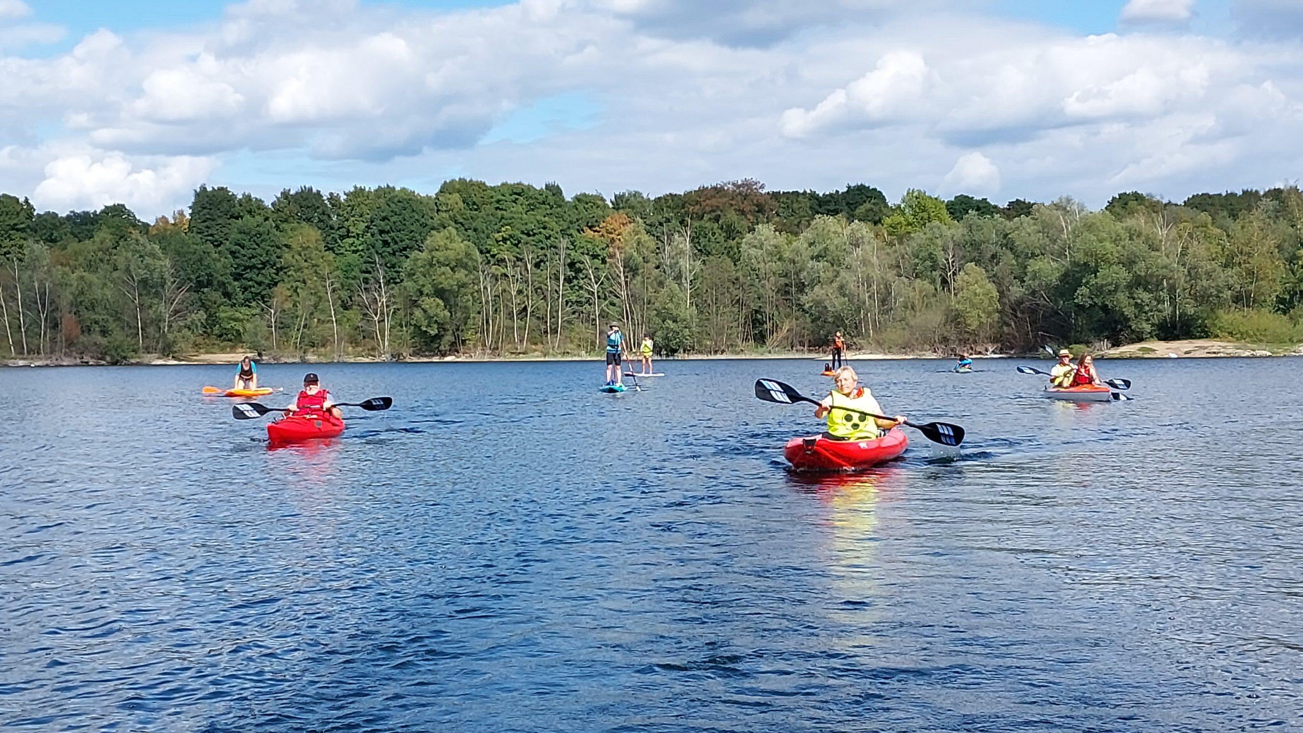 Mehrere Paddlerinnen und Paddler mit Begleitpersonen in Kajaks und auf SUP-Boards auf einem von Wald umgebenen See.